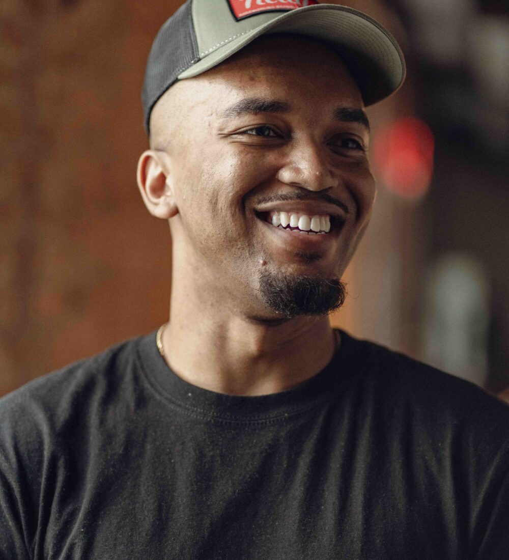 Man in black T-shirt and cap smiles indoors.