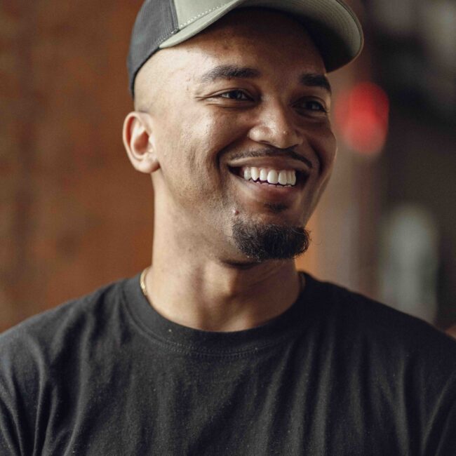 Man in black T-shirt and cap smiles indoors.