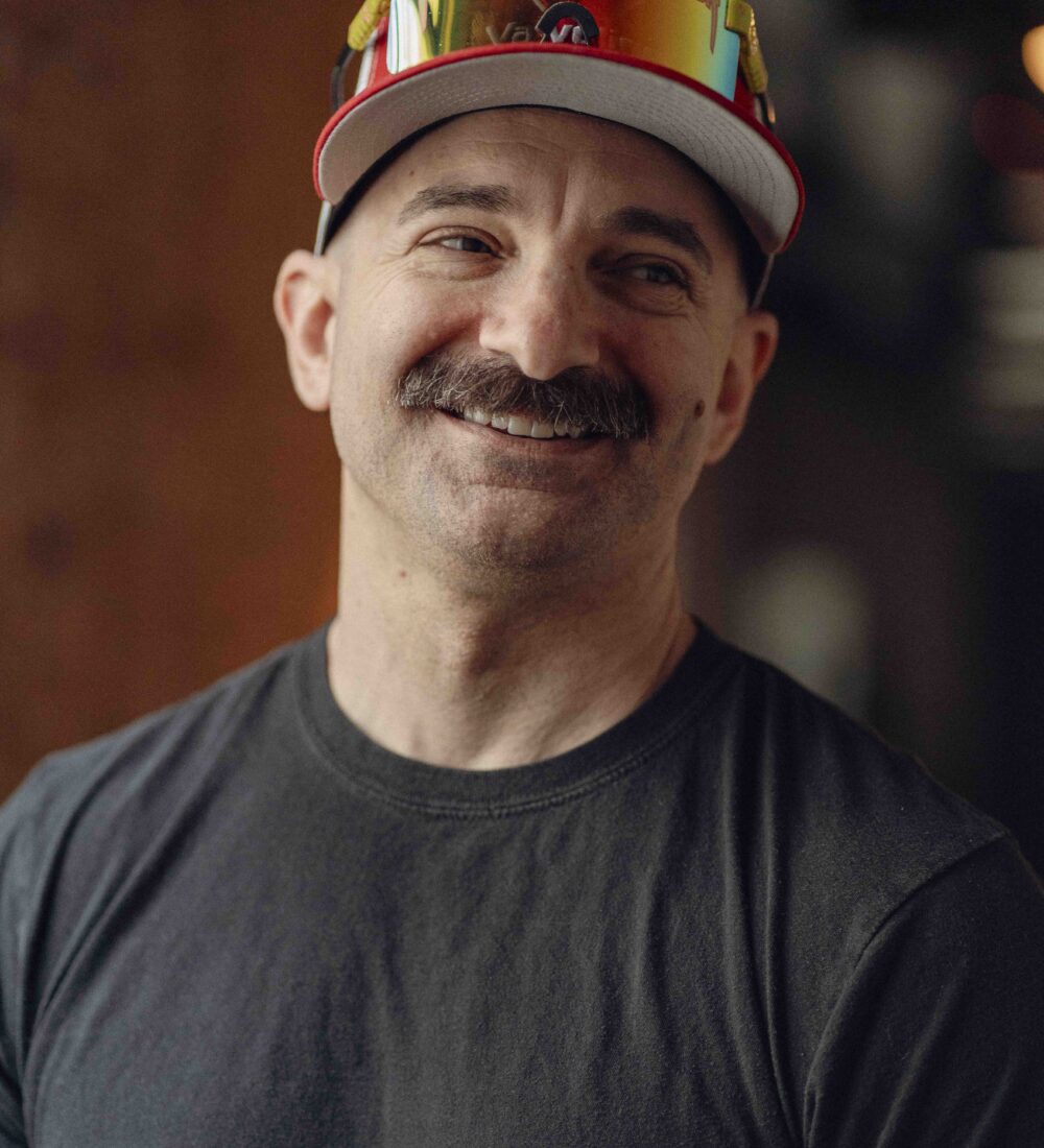 Smiling mustached man in black shirt, sunglasses on hat.