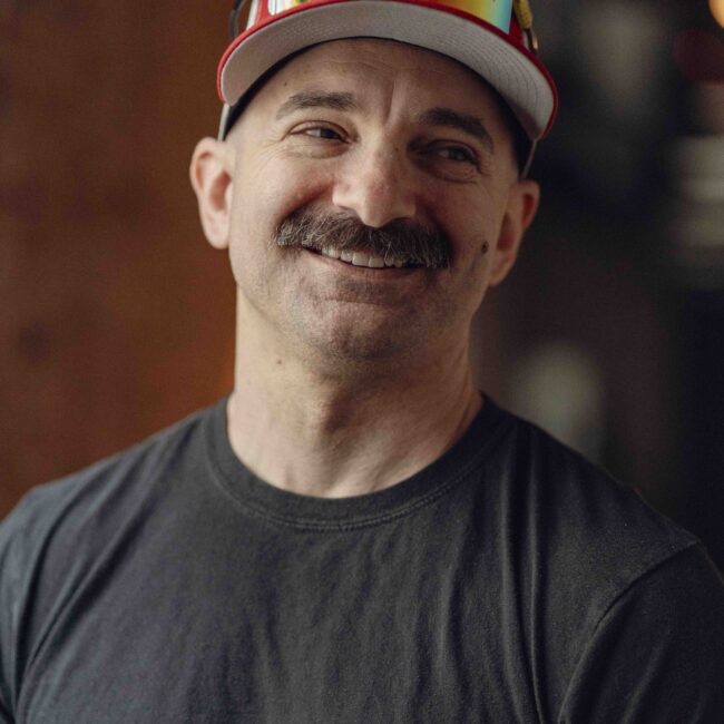 Smiling mustached man in black shirt, sunglasses on hat.