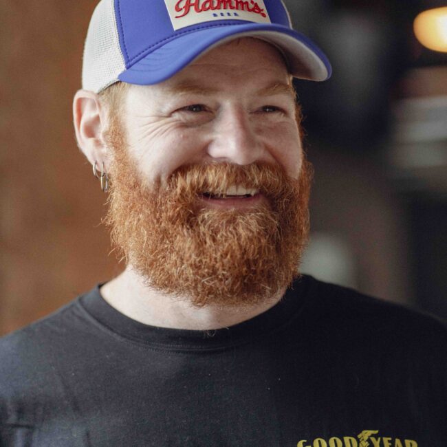 A bearded man in a cap and black Goodyear T-shirt smiles.