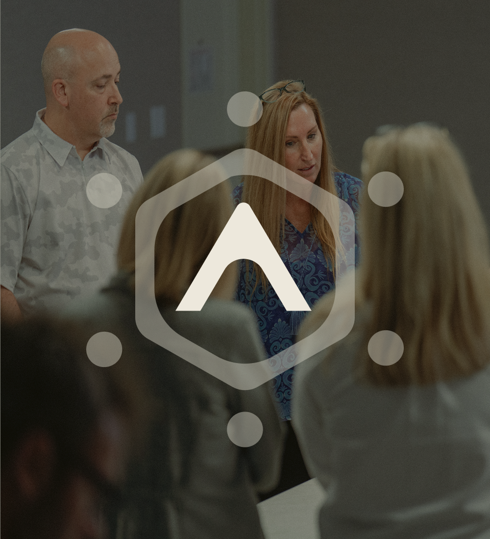 Two people address a seated group with hexagon and overlay.