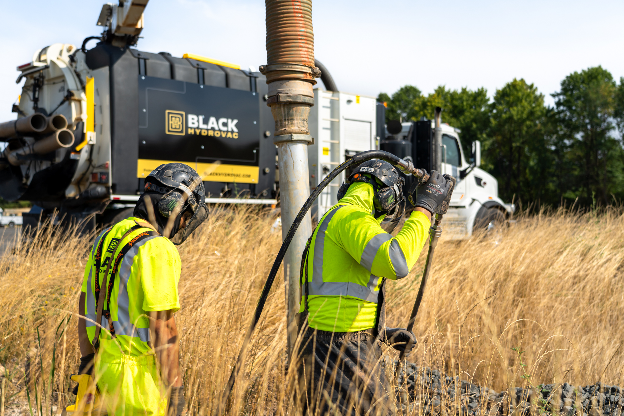 Workers in bright gear excavate near black hydrovac truck.