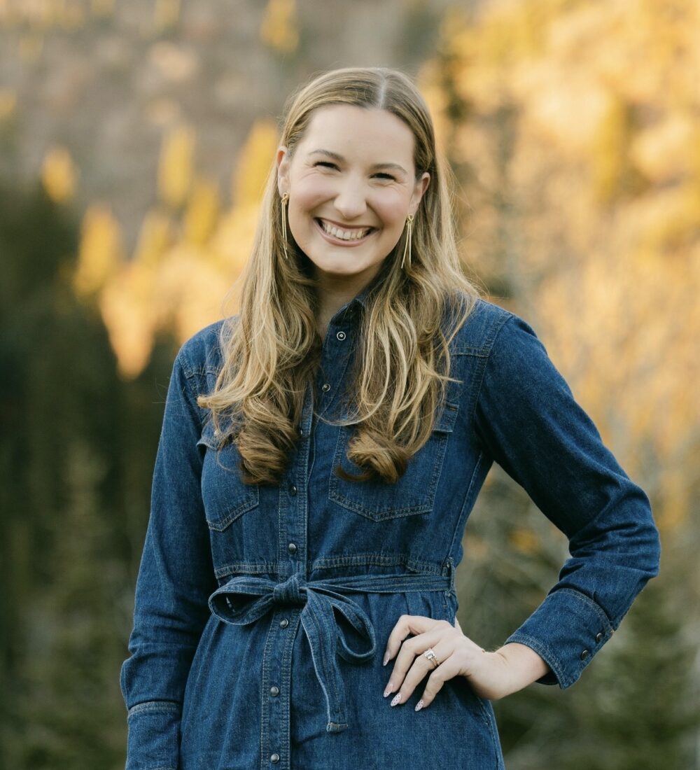 Blonde woman in denim dress smiles outdoors by autumn trees.