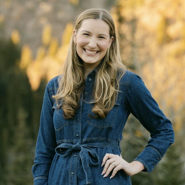 Blonde woman in denim dress smiles outdoors by autumn trees.