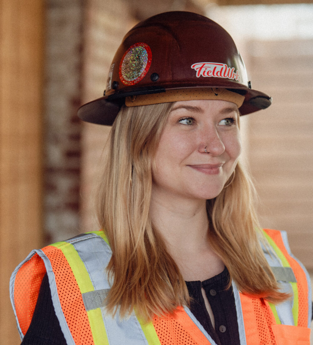 Woman with blonde hair in a hard hat and safety vest smiles outside