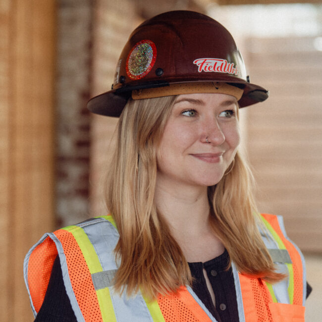 Woman with blonde hair in a hard hat and safety vest smiles outside