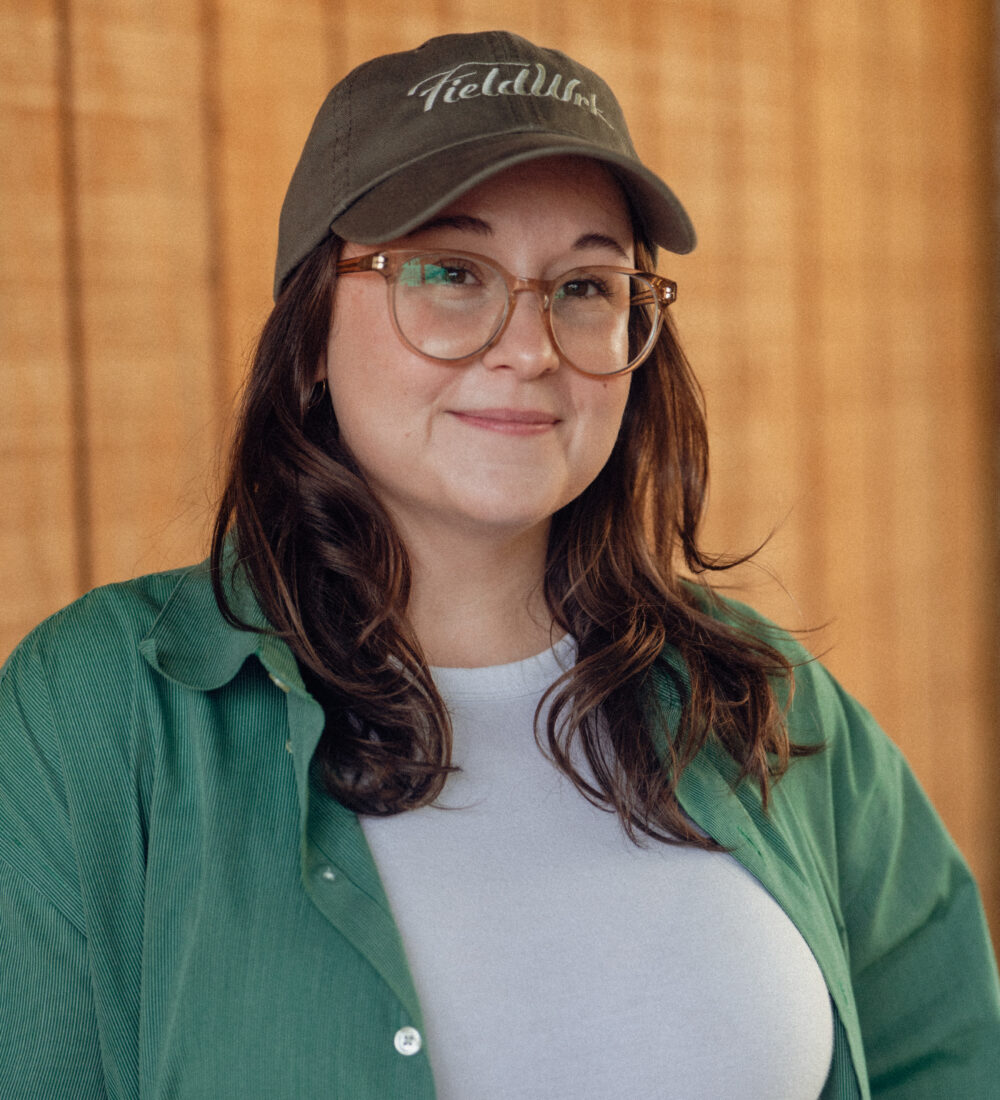 Woman in glasses, green shirt, and cap smiles by wood background.