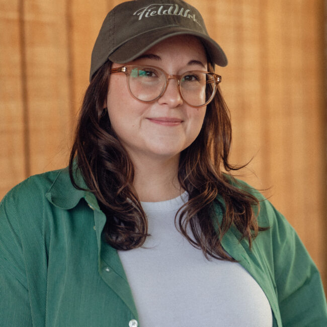 Woman in glasses, green shirt, and cap smiles by wood background.