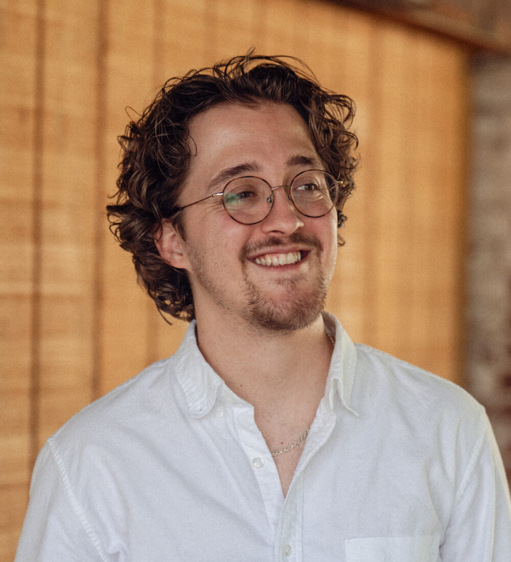 man in glasses and a white shirt smiles in front of a wood background