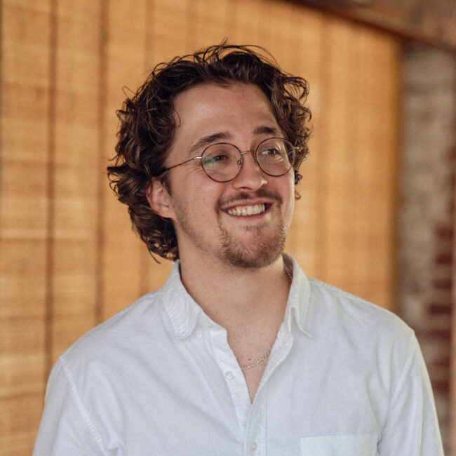 man in glasses and a white shirt smiles in front of a wood background