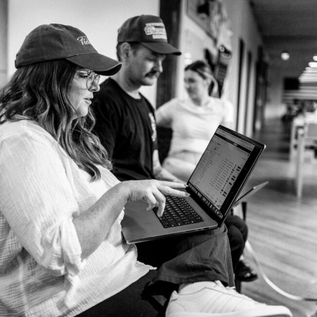 Woman in cap uses laptop; two others sit nearby, indoors.
