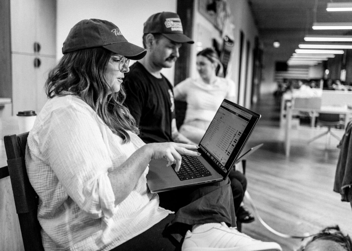 Woman in cap uses laptop; two others sit nearby, indoors.