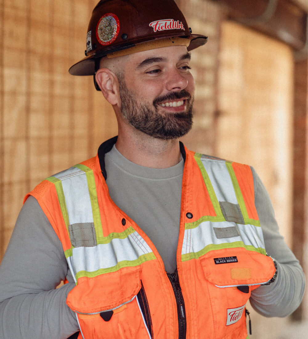 Man in a hard hat and orange safety vest smiles outside