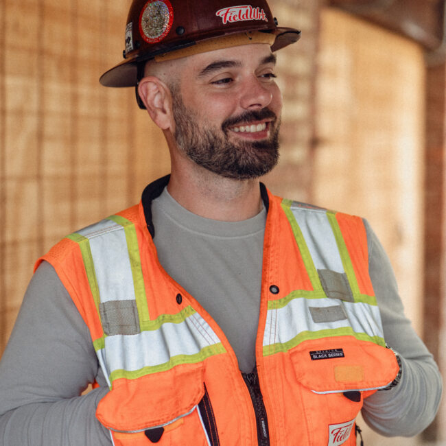 Man in a hard hat and orange safety vest smiles outside