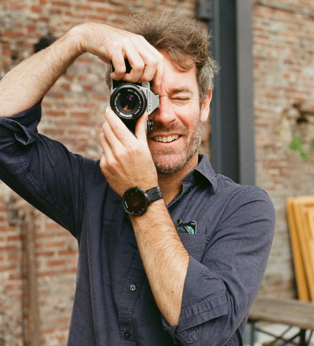 Bearded man in dark shirt smiles, taking photo with film camera.