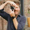 Bearded man in dark shirt smiles, taking photo with film camera.