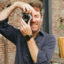 Bearded man in dark shirt smiles, taking photo with film camera.