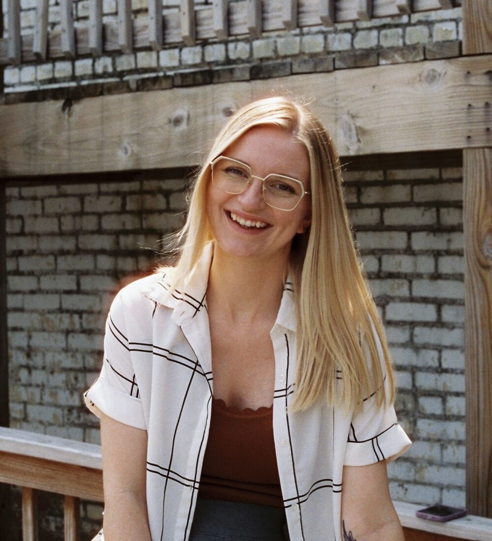 Smiling blonde woman in glasses sits by brick wall and railing.