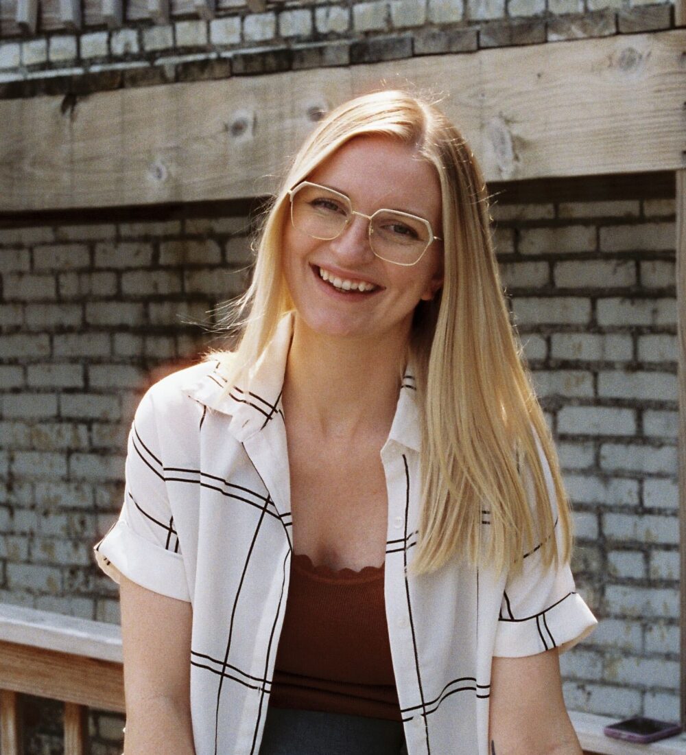Smiling blonde woman in glasses sits by brick wall and railing.