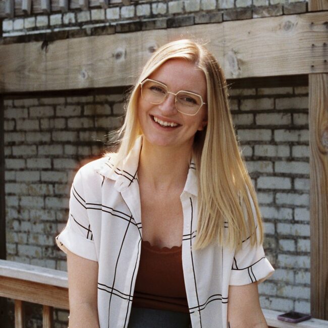 Smiling blonde woman in glasses sits by brick wall and railing.