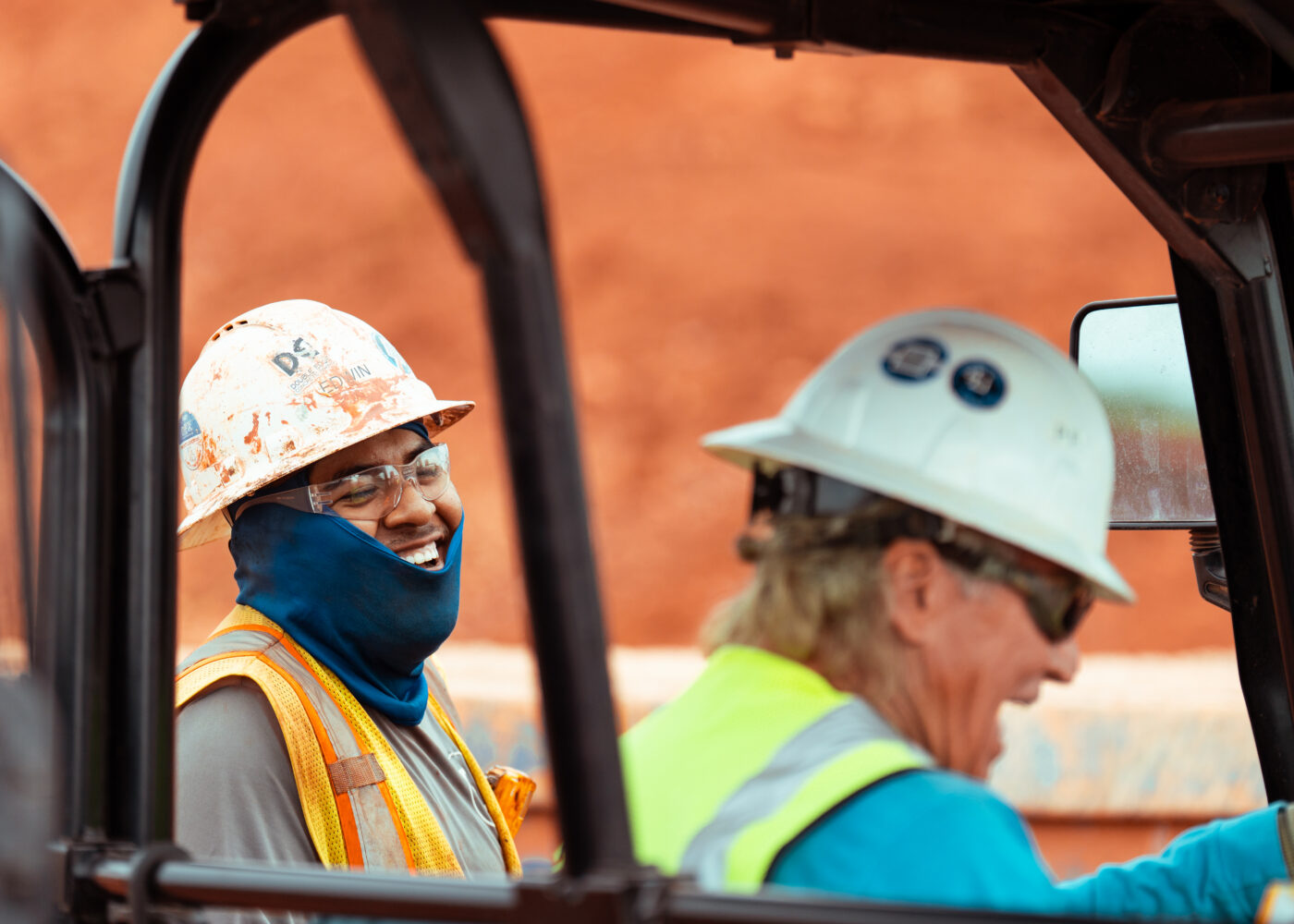 Two construction workers in safety gear by a vehicle; one smiles.
