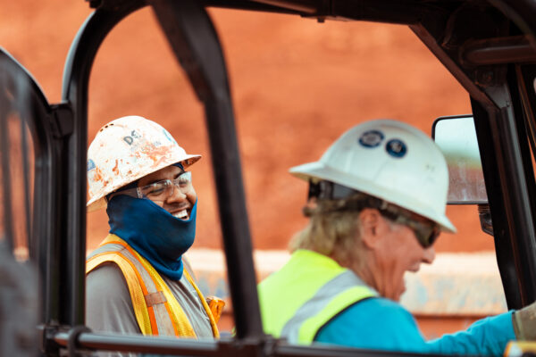 Two construction workers in safety gear by a vehicle; one smiles.