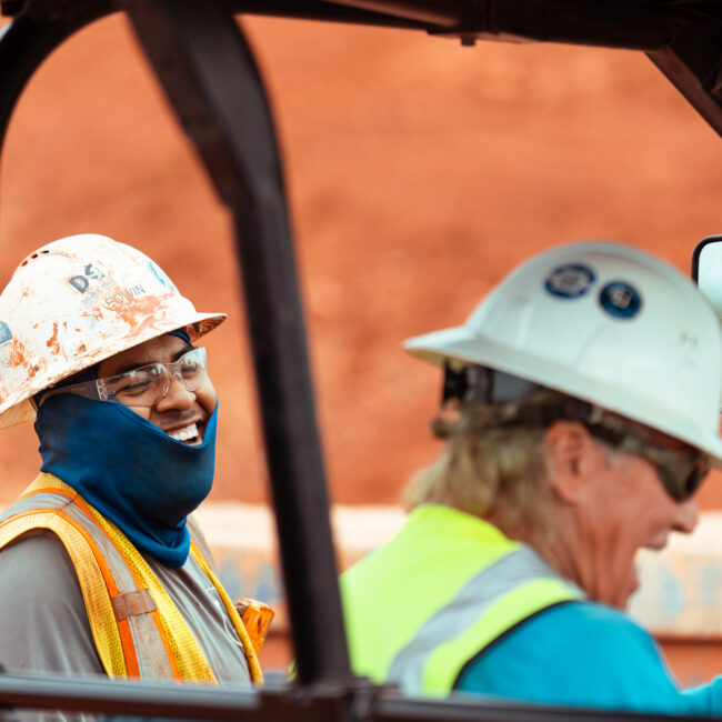 Two construction workers in safety gear by a vehicle; one smiles.