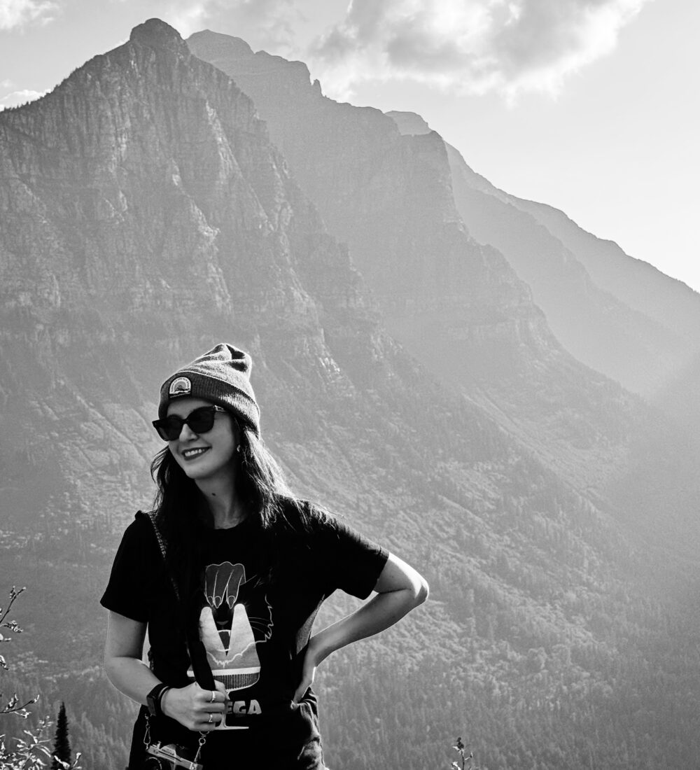 Woman in beanie and sunglasses holds camera by mountains.
