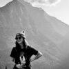 Woman in beanie and sunglasses holds camera by mountains.