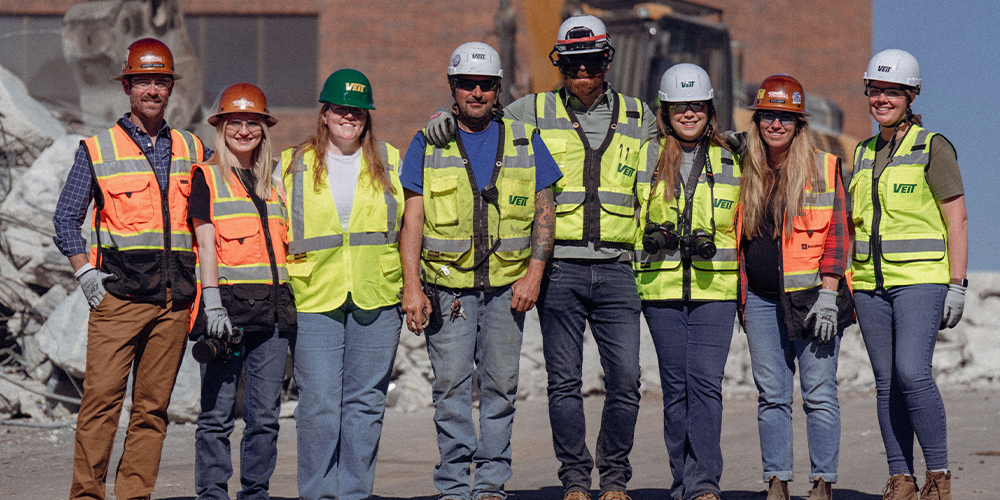 Eight workers in safety gear stand at a rubble-filled site.