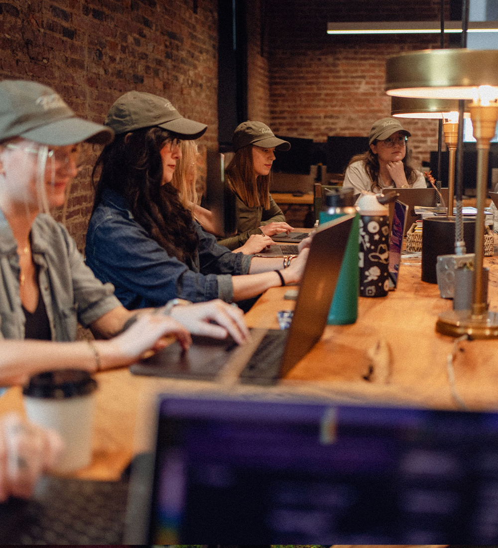 Four people in hats work on laptops around a table.