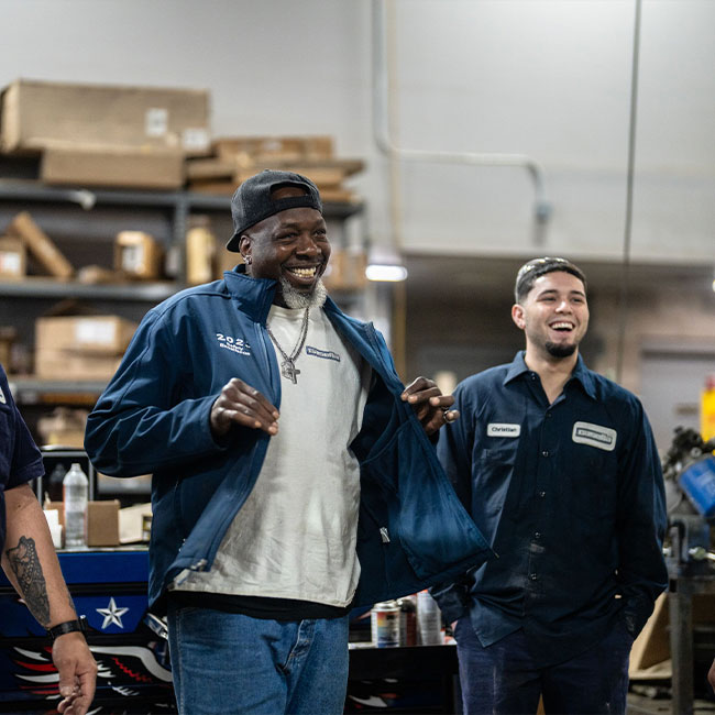 Two men in work uniforms smile in an industrial workshop.
