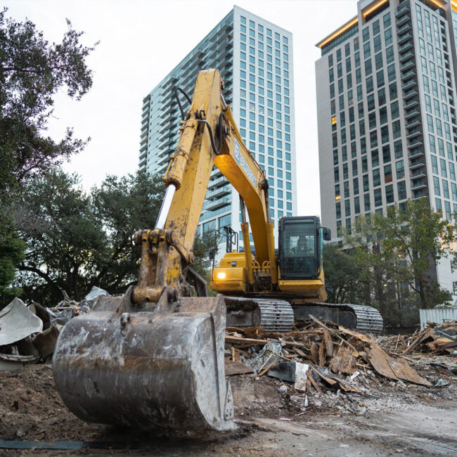 Yellow excavator at construction site, debris and buildings behind.