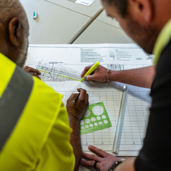 Two people in hi-vis shirts measure and mark architectural plans.