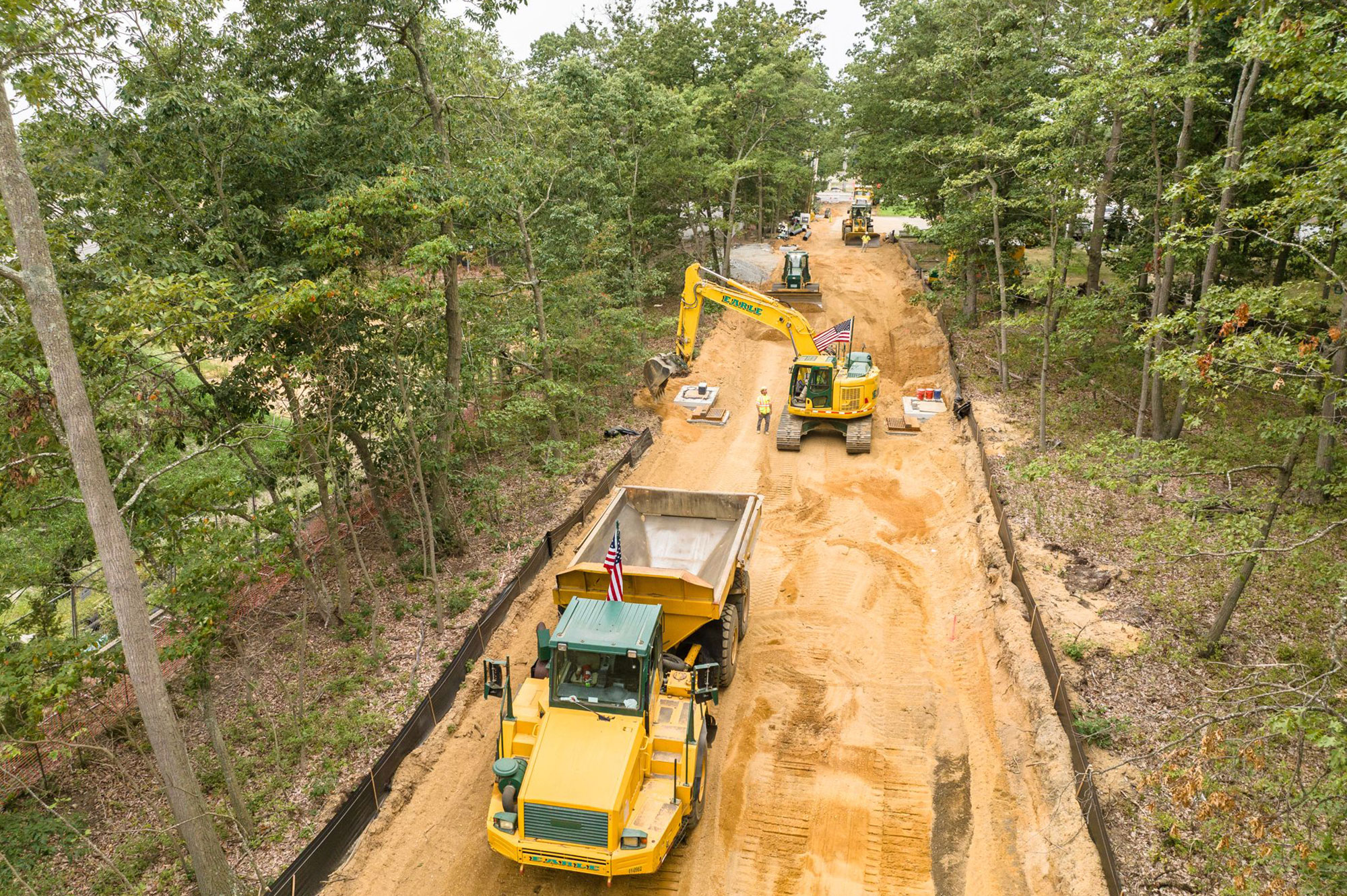 Excavators and dump trucks work on a dirt road in woods.