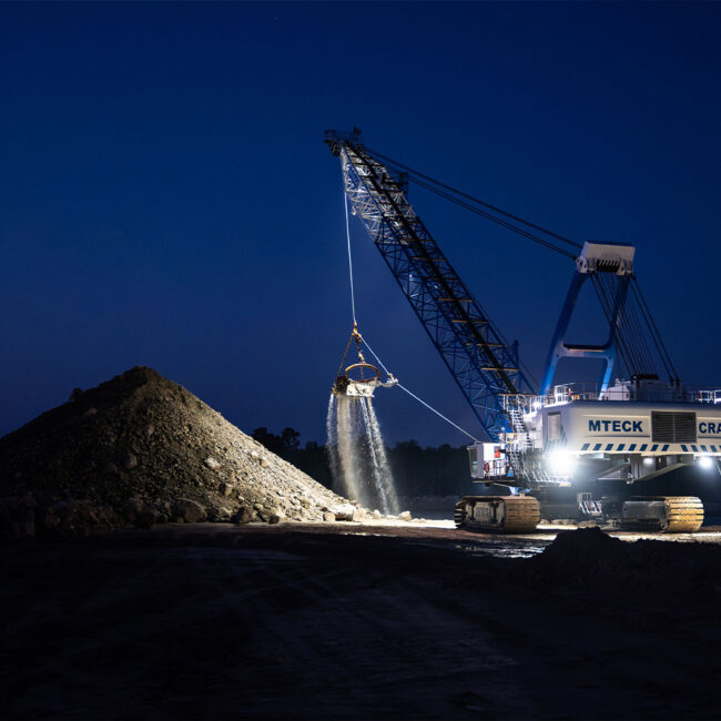 Excavator with bright lights moves earth onto a pile at night.