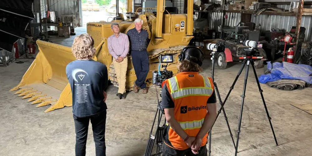 Two people with a yellow bulldozer and camera crew in garage.