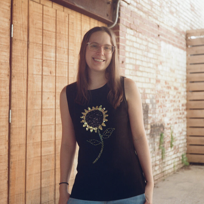 Person with long hair and glasses smiles outside near a wall.