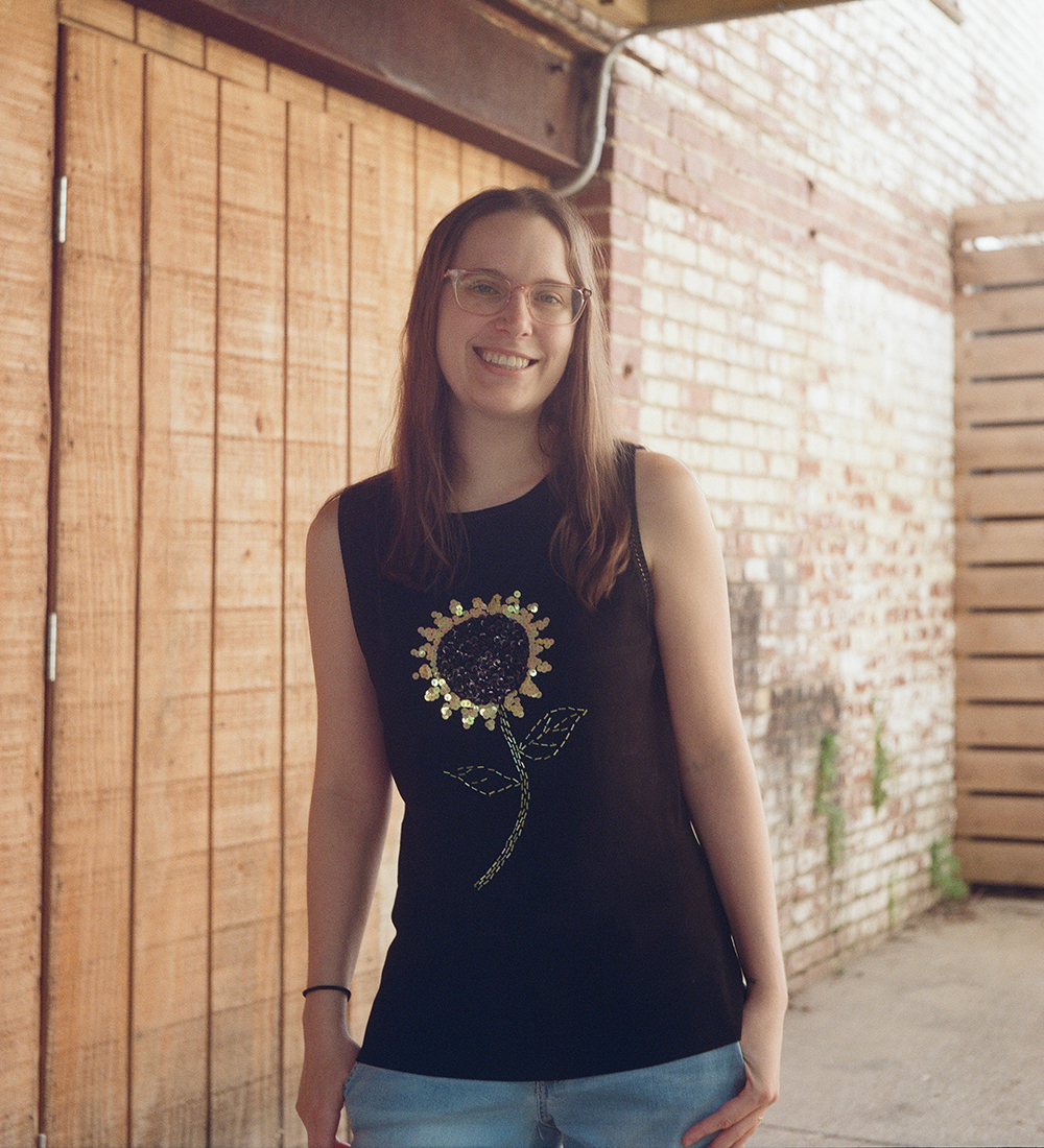 Person with long hair and glasses smiles outside near a wall.