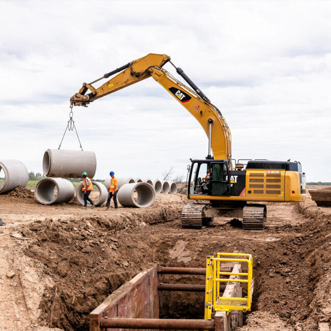Excavator lifts concrete pipe as two workers guide it.