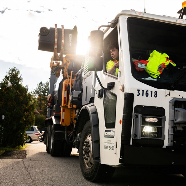 Sanitation worker drives white garbage truck on sunny street.