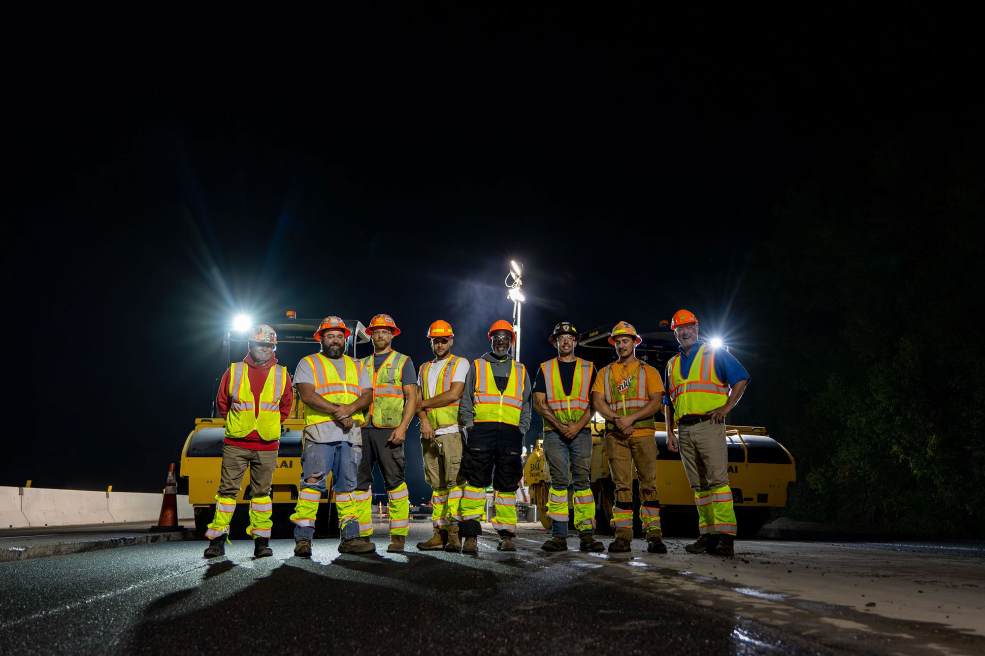 Seven construction workers in gear stand on a road at night.