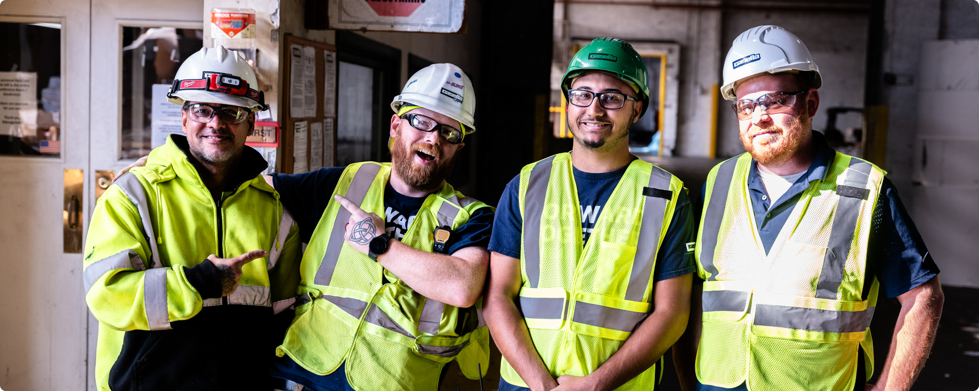Four smiling construction workers in safety vests stand indoors.
