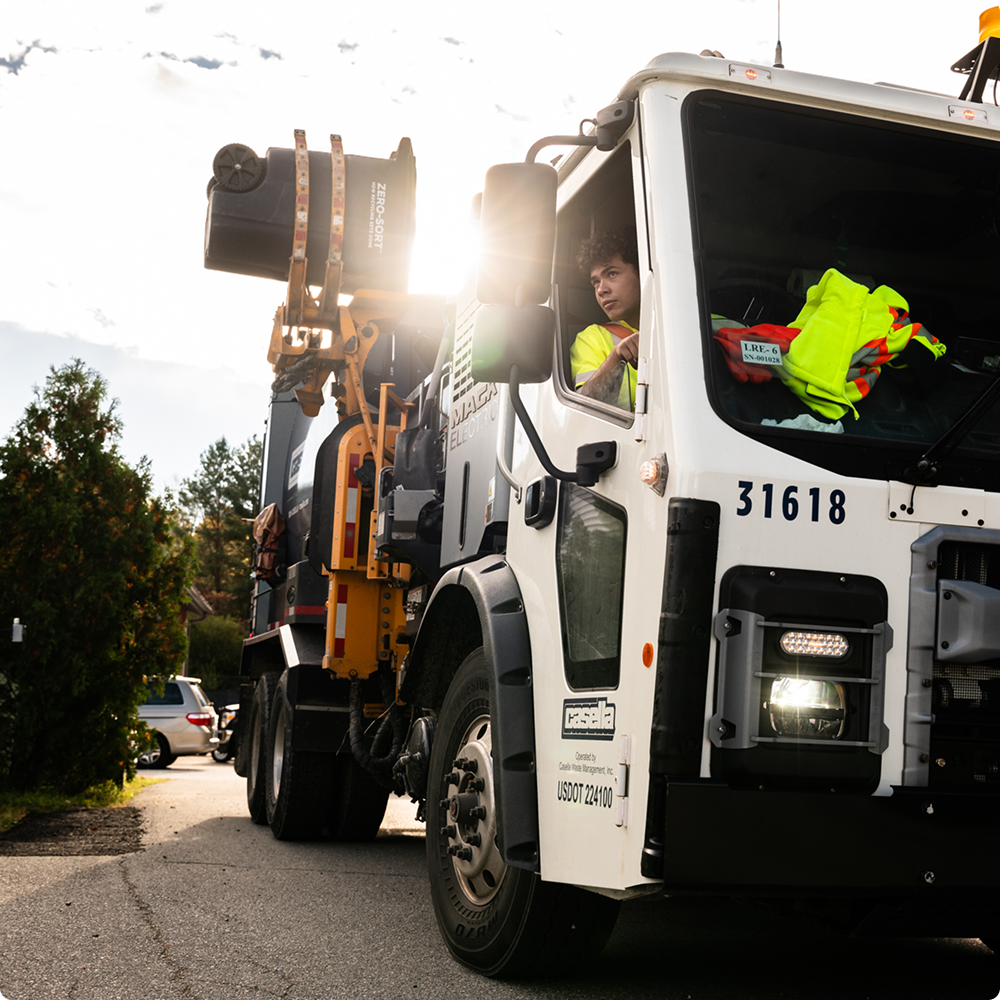 Worker drives white utility truck with equipment on sunny street.