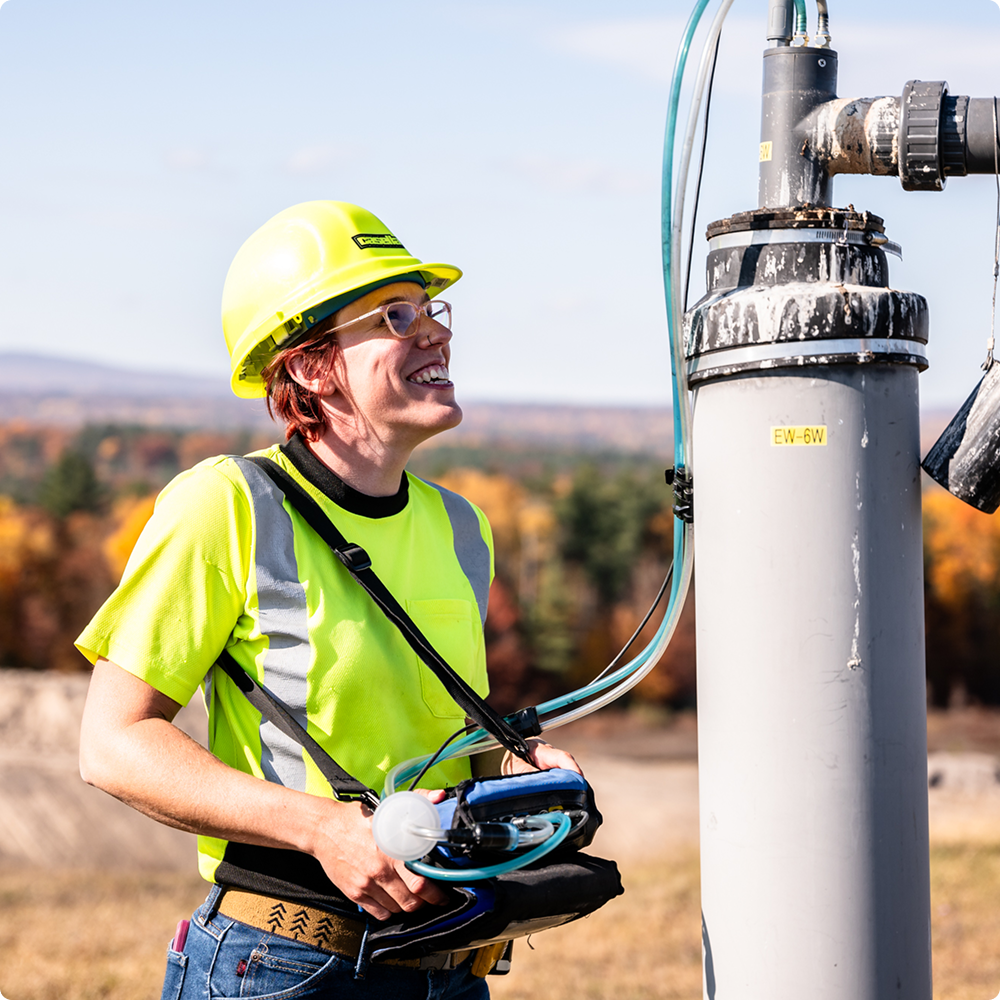 Worker in yellow hard hat inspects outdoor equipment by large pipe.