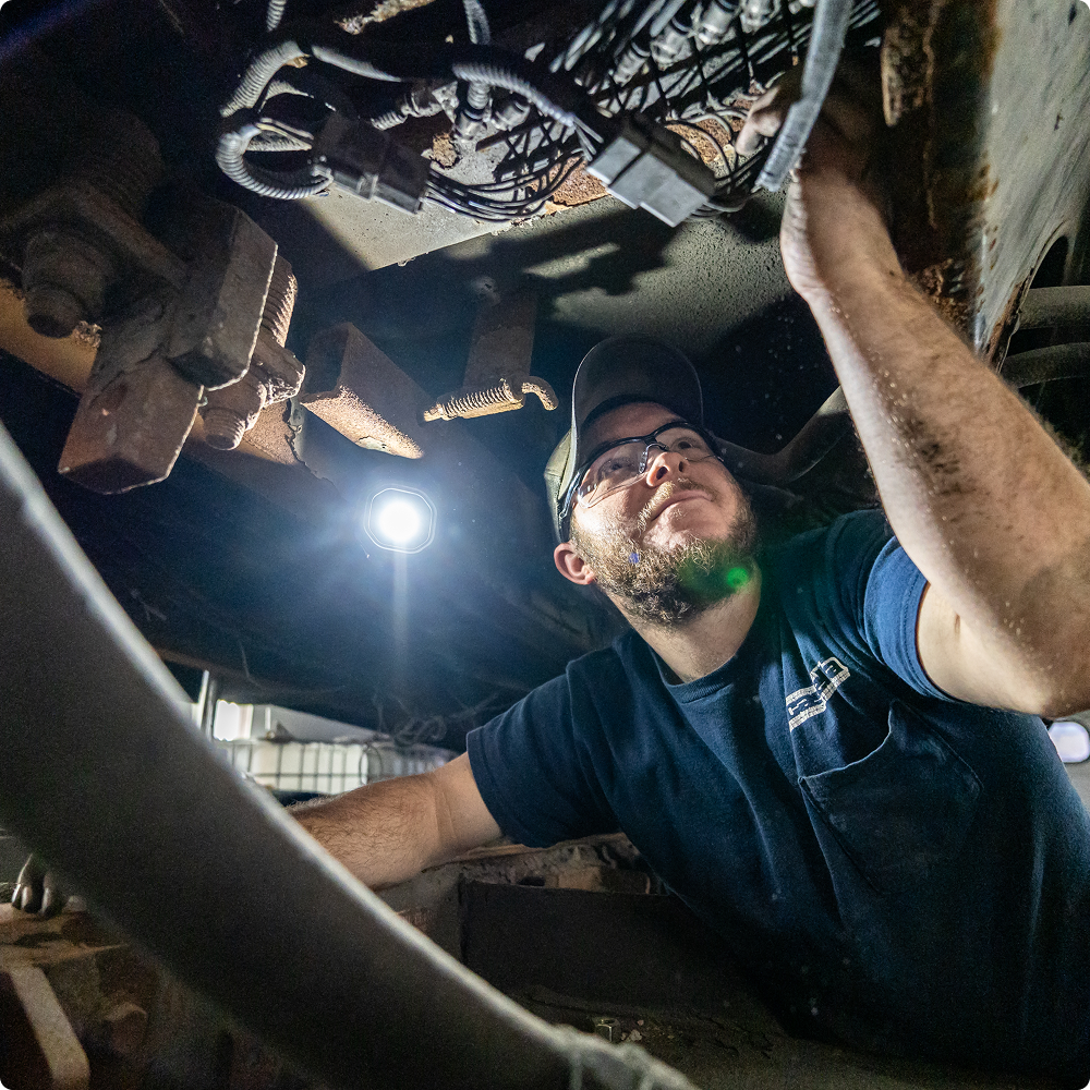 Person in cap and glasses works on wiring under vehicle.
