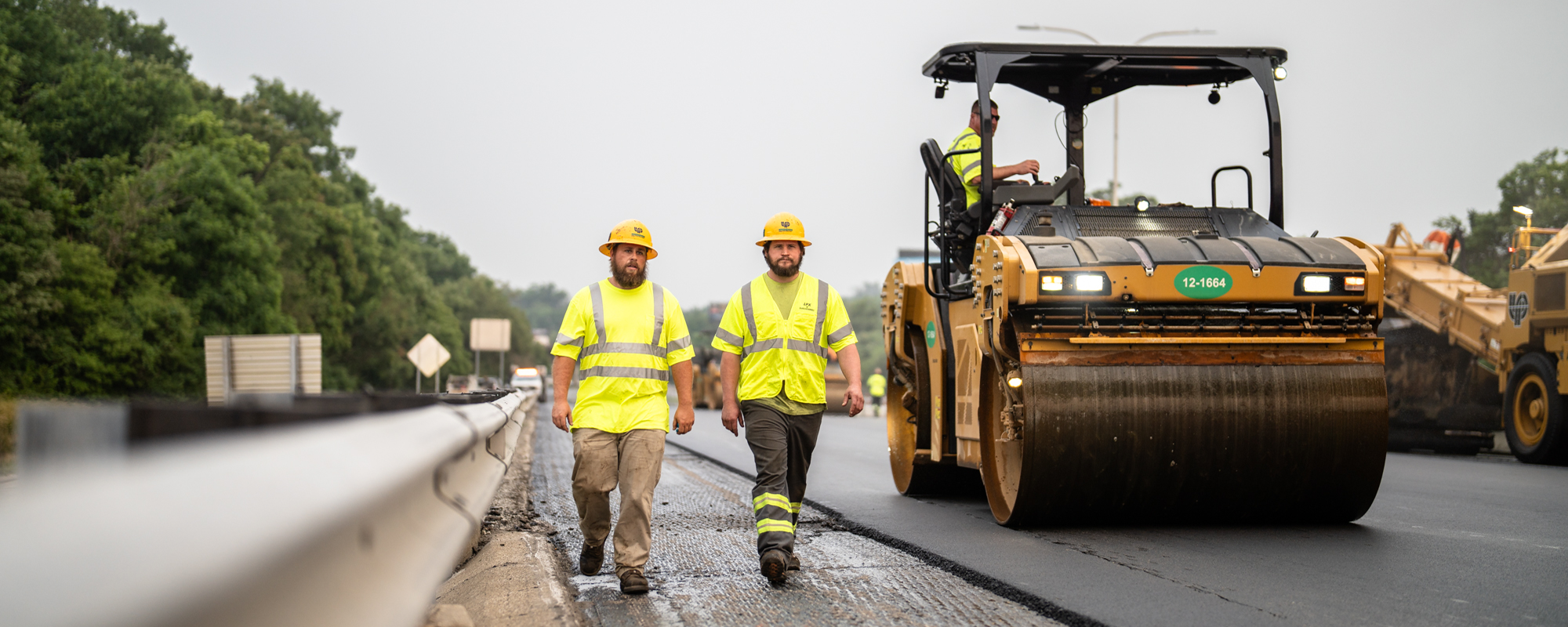 Two workers walk on highway, another operates road roller.