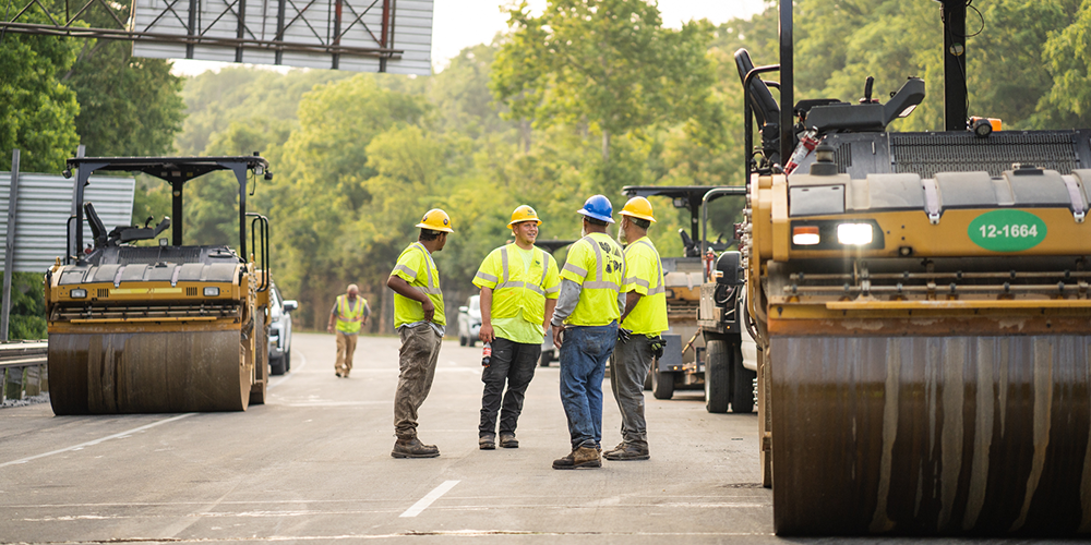 Four workers in safety gear stand on a road between paving machines.
