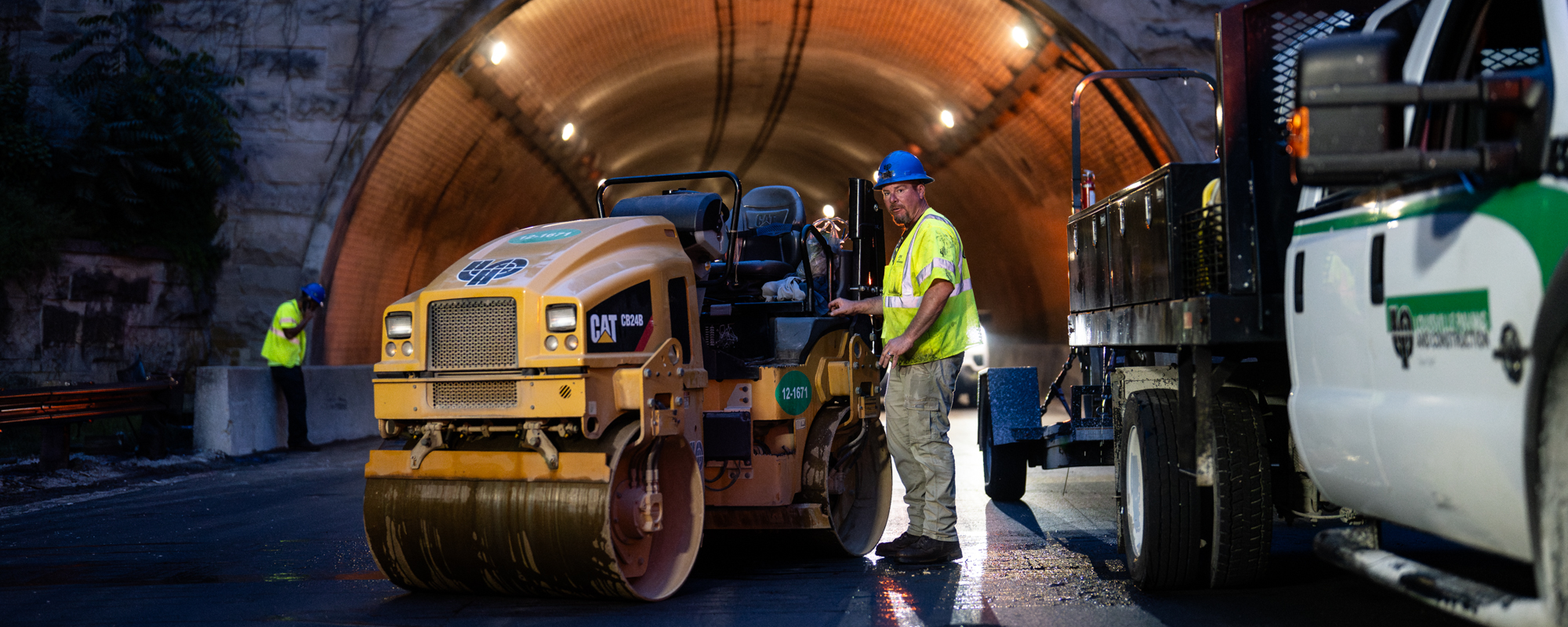 Construction worker drives road roller at night near tunnel.